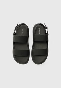 Svarte sandaler laget av syntetisk materiale har to brede stropper og en justerbar ankelrem, samt en flat såle og et minimalistisk design.
