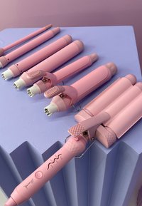 Ensemble d'outils à boucler roses, de tailles et de formes variées, avec des poignées texturées et des embouts blancs, disposés sur une surface violette.
