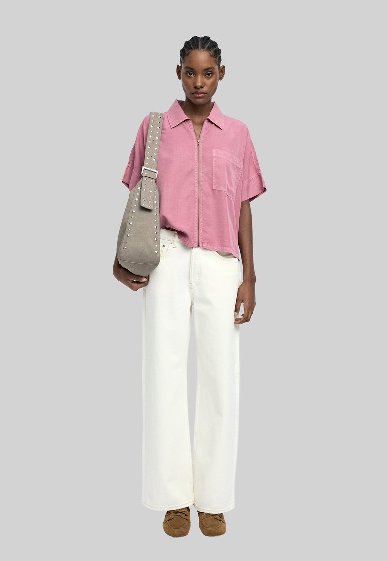 Jasnoróżowa koszula z kołnierzykiem i krótkim rękawem, białe szerokie dżinsy i brązowe zamszowe buty. Torebka z ćwiekami na pasku. Neutralne tło.