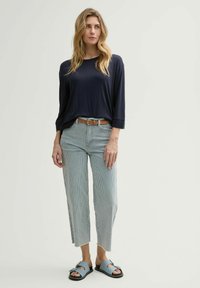 Navy lange mouwen top, licht gestreepte wijde jeans met franjes, en blauwe slidesandalen met een gesp-accent. Bruine riem inbegrepen.