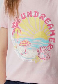 Roze katoenen t-shirt met een kleurrijke strandscène featuring palmbomen, zon en schelpenafbeelding in glitter en pastelkleuren. Inclusief de tekst "SUNDREAMER".