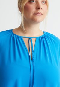 Blauweblouse van gladde, lichte stof met een ronde halslijn, een keyhole-detail aan de voorkant en geplooide accenten bij de hals.