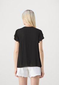 Svart bluse med volanger og korte ermer, laget av strukturert stoff, med rund utringning og løs passform, båret over hvite frayed shorts.