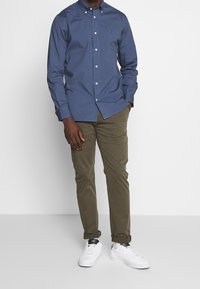 Camicia blu con bottoni, colletto, maniche lunghe e un piccolo logo sul petto, abbinata a pantaloni verde oliva e sneakers bianche.