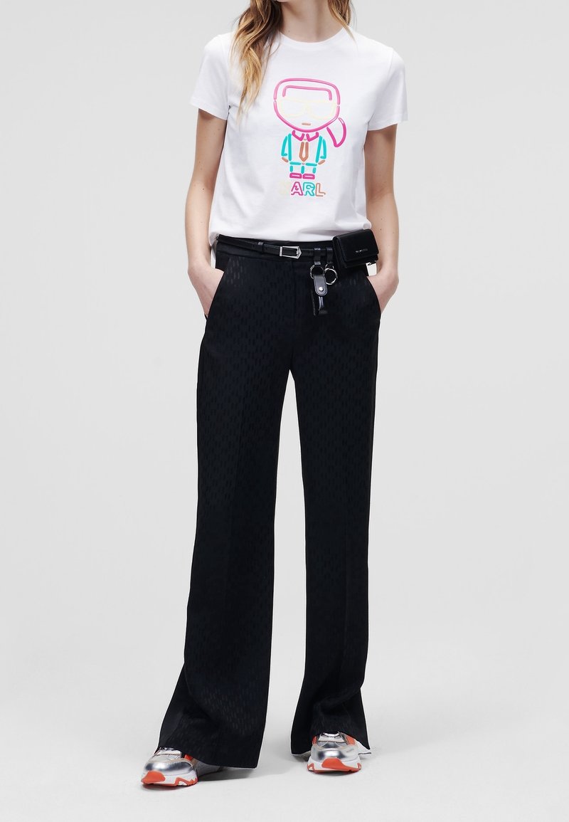 Camiseta blanca con un gráfico de dibujos animados colorido y texto, combinada con pantalones anchos negros con estampado y zapatillas plateadas con detalles en naranja.
