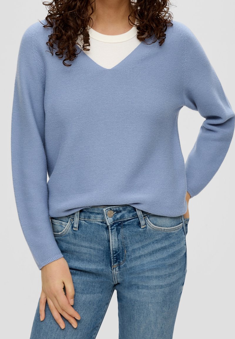 Lichtblauwe gebreide trui met een V-hals en lange mouwen, gecombineerd met lichtblauwe denim jeans. De textuur is ribbelig en zacht.