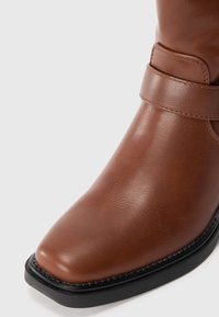 Gros plan sur une botte en cuir marron avec un détail de sangle et une semelle noire sur fond blanc uni.