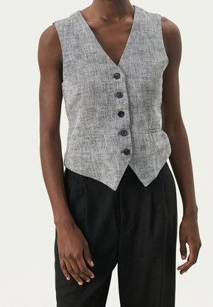 Veste grise sans manches boutonnée avec deux poches avant portée avec un pantalon noir sur une personne à la peau foncée sur un fond uni.