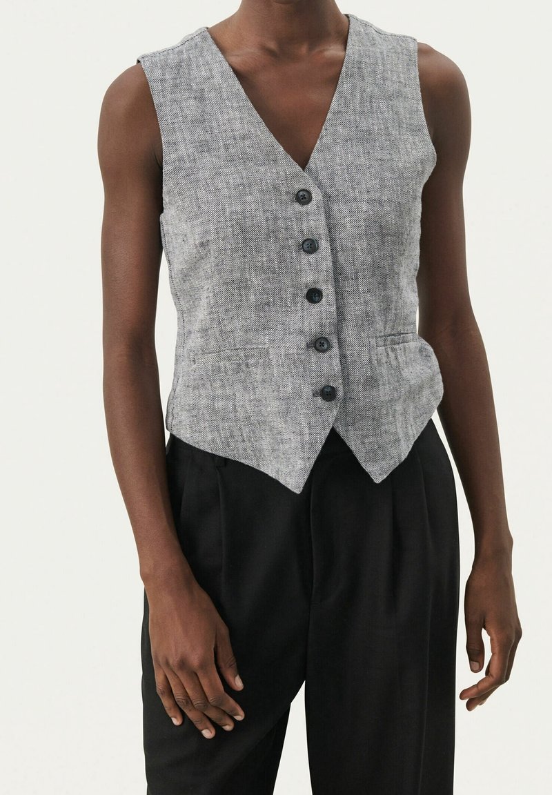 Veste grise sans manches boutonnée avec deux poches avant portée avec un pantalon noir sur une personne à la peau foncée sur un fond uni.