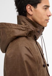 Brown waterproof jacket with a high collar and hood. Features drawstring closure and smooth texture. Side profile view highlights sleek design.