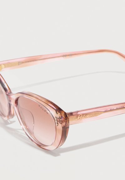 Ροζ διάφανα τετράγωνα γυαλιά ηλίου με φακούς με διαβάθμιση χρώματος και λεπτούς βραχίονες που ακουμπούν σε λευκή επιφάνεια.