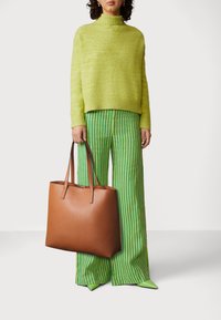 Borsa a tote in pelle marrone con manici lunghi, caratterizzata da una texture liscia e hardware minimale. Il modello indossa un maglione verde lime e pantaloni a righe.