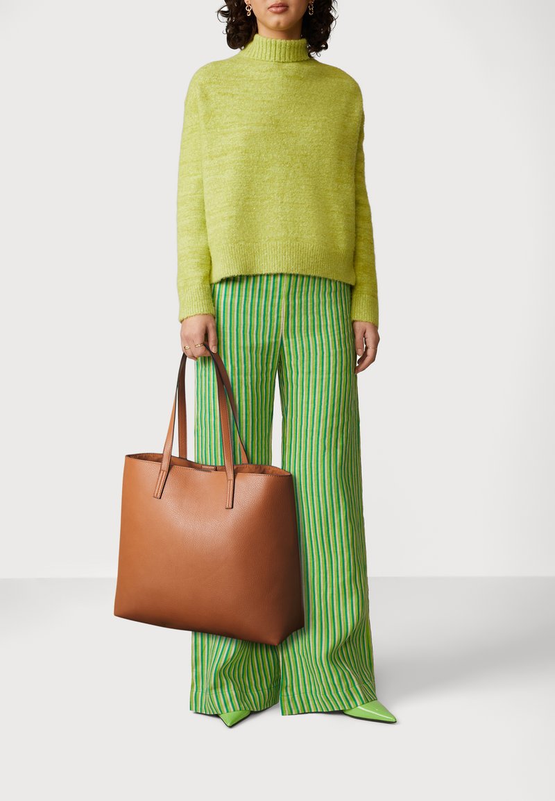 Borsa a tote in pelle marrone con manici lunghi, caratterizzata da una texture liscia e hardware minimale. Il modello indossa un maglione verde lime e pantaloni a righe.