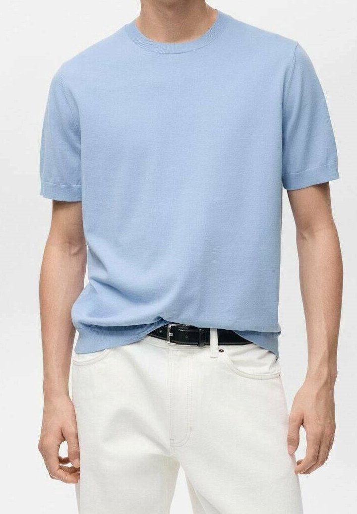 Homme portant une chemise en maille bleu clair à manches courtes rentrée dans un pantalon blanc avec une ceinture noire, debout devant un fond uni.