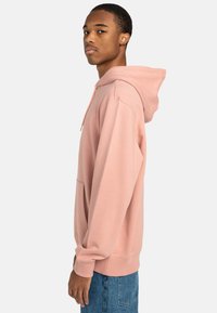 Felpa rosa con cappuccio, dotata di tasca a marsupio e polsini a costine. Il tessuto sembra morbido e texturizzato, indossato da un modello di lato.
