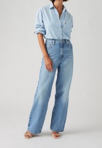 Femme portant une chemise en denim bleu clair à manches retroussées, un jean bleu large taille haute et des talons ouverts beige, debout devant un fond uni.
