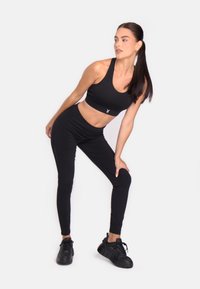 Ženska v črnem športnem modrčku, legginseh in športnih čevljih, ki se nagiba naprej z eno roko na kolenu, ob beli podlagi.