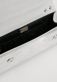 Zilverkleurige metallic handtas met een zachte textuur, heeft een zwart stoffen interieur, geplooide voering en een klein logoplaatje aan de binnenkant.