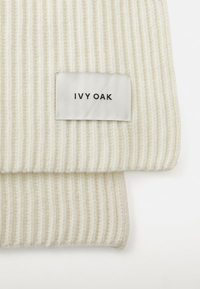 Balta ribuota megztinė skara su audinio etikete, kurioje juodu tekstu užrašyta "IVY OAK". Audinys yra minkštas ir storas, pabrėžiantis megztą raštą.