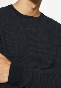Schwarzer strukturierter Pullover mit einem gerippten Ausschnitt und Bündchen, der ein gewebtes Muster und eine lockere Passform aufweist.