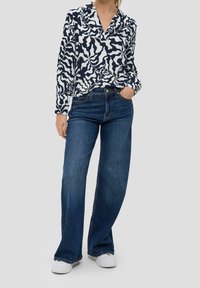 Kvinna som bär en marinblå och vit blus med abstrakt mönster, mörkblå vida jeans och vita sneakers, stående med ena handen i fickan.