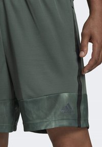 Närbild av en person som bär gröna Adidas-shorts med svarta sidosträck och logotyp nära fållen, stående med handen avslappnad vid sidan.