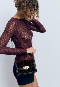 Donna con capelli ricci che indossa un top in pizzo trasparente bordeaux con maniche lunghe e una gonna nera, mentre tiene una borsa con stampa leopardata e tracolla in oro.