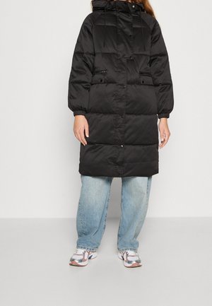 Persoon draagt een lange zwarte gewatteerde jas met capuchon, lichtblauwe spijkerbroek en witte sneakers tegen een effen grijze achtergrond.