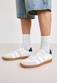 Sneakers bianchi con accenti verde sul tallone, suole in gomma color beige e strisce laterali grigio chiaro. Indossati con calze bianche a coste e pantaloni shorts in denim.