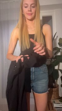 Crna tkanina odjeće držana u rukama, u kombinaciji s trapericama. Svijetla plava kosa, jednostavna ogrlica i sobno bilje u pozadini.