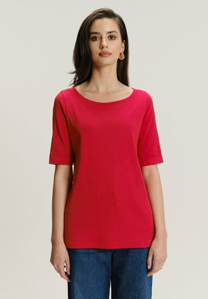 Kobieta z ciemnymi włosami, nosząca złote kolczyki w kształcie kół, czerwoną koszulkę z krótkim rękawem i niebieskie dżinsy, stojąca na jednolitym jasnym tle.