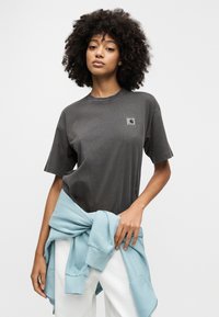 Γυναίκα με σγουρά μαλλιά φοράει σκουρόχρωμο γκρι μπλουζάκι και άσπρο παντελόνι με ένα ανοιχτό μπλε φούτερ δεμένο γύρω από τη μέση της, στέκεται μπροστά σε λευκό φόντο.