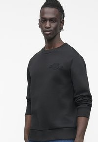 Mladý muž s krátkými dreadlocky, oblečený v černé mikině s kulatým výstřihem a jemným logem na hrudi, stojí před jednobarevným bílým pozadím.