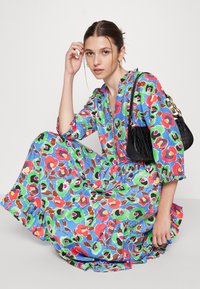 Vestido maxi com padrão floral em azul, apresentando acabamentos em vermelho, verde e castanho. A modelo segura uma pequena bolsa preta com acabamento texturizado.