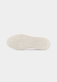 Semelle de chaussure en caoutchouc blanche avec motifs en zigzag et circulaires, talon texturé, et logo "GUESS" embossé près du talon.