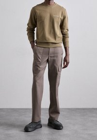 Man klädd i en olivgrön sweatshirt och taupe cargo-byxor med svarta skor, ståendes på en grått golv mot en enkel ljus vägg.