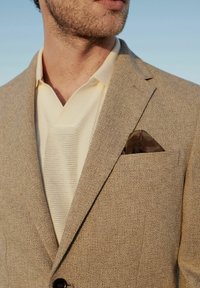 Blazer de textura marrón sobre un polo de punto crema, con un pañuelo de bolsillo de patrón marrón en el bolsillo del pecho.