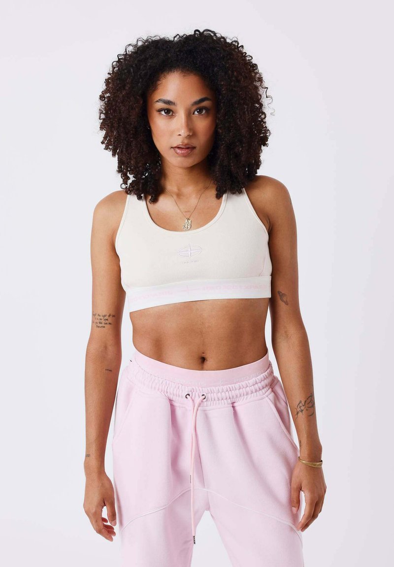 Brassière de sport crème claire avec une coupe ajustée et un logo violet ; associée à un pantalon de jogging rose ample avec une ceinture élastique.