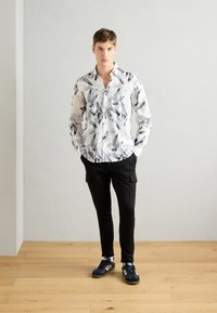Hvit skjorte med svart bladmønster, knappefeste foran, lange ermer. Kombination med svarte cargo-bukser og svarte sneakers. Bakgrunn med tregulv.