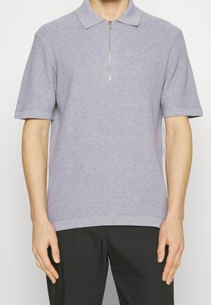 Mann trägt ein hellgraues kurzärmliges Strick-Polo mit Frontreißverschluss und schwarze Hose, steht vor einem einfarbigen weißen Hintergrund.