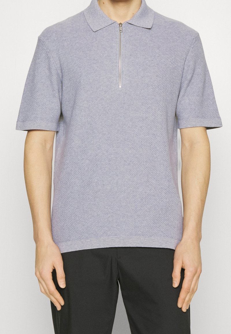 Homme portant un polo en maille gris clair à manches courtes avec une fermeture éclair à l'avant et un pantalon noir, debout devant un fond blanc uni.