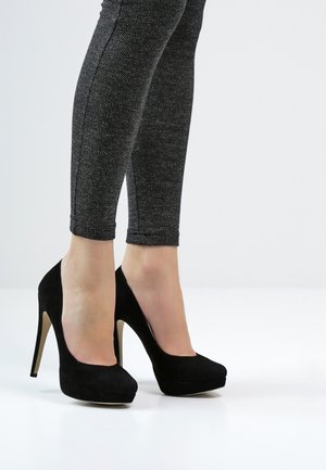 Chaussures à talons hauts en daim noir avec un bout rond et une plateforme, dotées d'un talon aiguille. Portées avec des leggings texturés sombres.