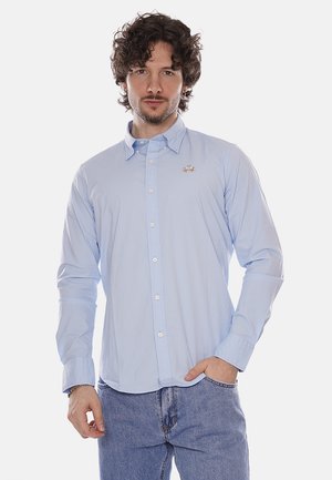 Uomo con capelli ricci che indossa una camicia azzurra con bottoni e jeans blu, in piedi con una mano in tasca su sfondo bianco.