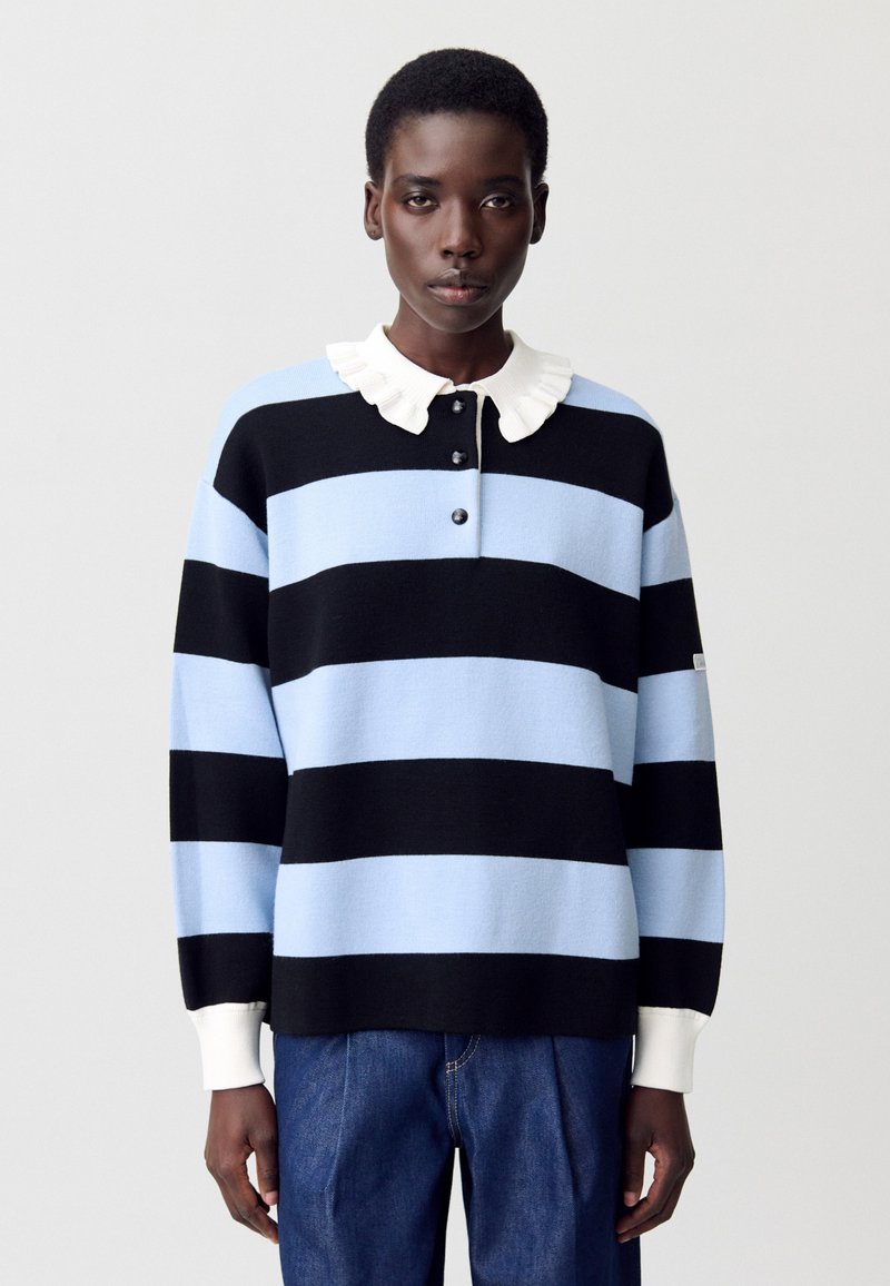 Suéter a rayas negro y azul cielo con un collar blanco con volantes, que presenta tres botones en el cuello y puños de canalé. Llevado con jeans de mezclilla.