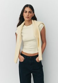 Krém színű, rövid ujjú, testhez simuló felső magas derekú fekete nadrággal párosítva. Egy rétegben egy puha, halvány sárga ujjatlan ruhadarabbal viselve.