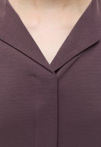 Gros plan d'une personne portant un t-shirt violet foncé à col en V avec un col et une couture verticale au centre devant.