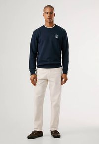 Sweatshirt azul-marinho com punhos e decote canelados, apresentando um logótipo circular no peito, combinado com calças bege claro e sapatos castanho escuro.