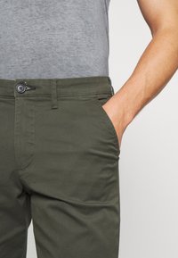 Khaki kalhoty s hladkým povrchem, slim fit a standardními kapsami. Pás má zapínání na knoflík a detaily s šňůrkou.