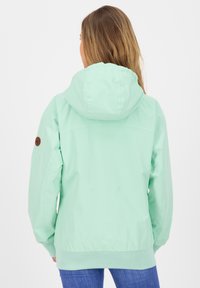 Mintgroene waterdichte jas met capuchon, elastische manchetten en een bruine embleem op de linker mouw. Glad stof, casual pasvorm.