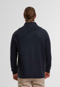 Marineblauer Strickpullover mit hohem, geripptem Kragen, langen Ärmeln und lockerer Passform. Weiche Textur mit dezentem Glanz, kombiniert mit beigen Hosen.
